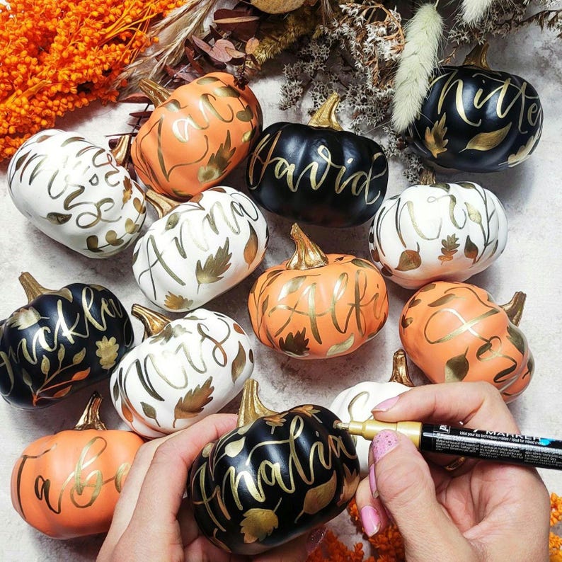 Pu&ograve; includere: Una collezione di piccole zucche decorative nere, bianche e arancioni, ciascuna con nomi scritti a mano e dettagli a foglia d'oro. Una mano tiene un pennarello dorato e scrive un nome su una zucca nera in primo piano.