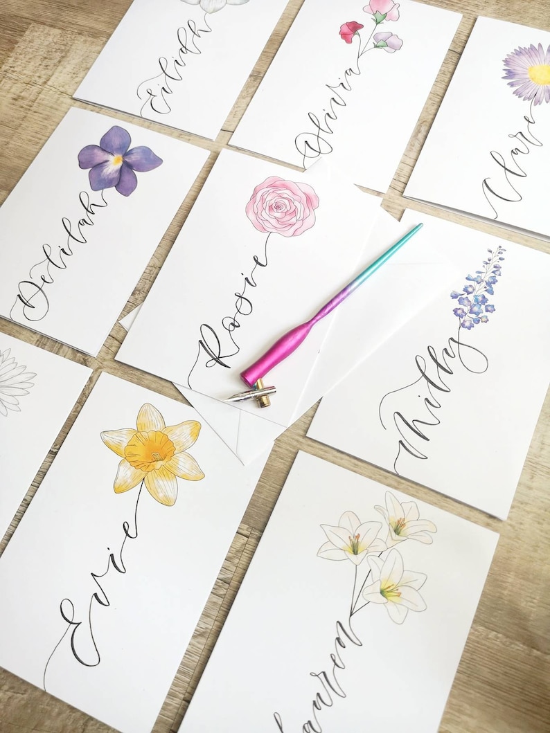 May include: A collection of white rectangular place cards featuring hand-lettered names in black ink paired with watercolor floral illustrations. A metallic pink and blue oblique calligraphy pen holder with a metal nib rests on the cards.