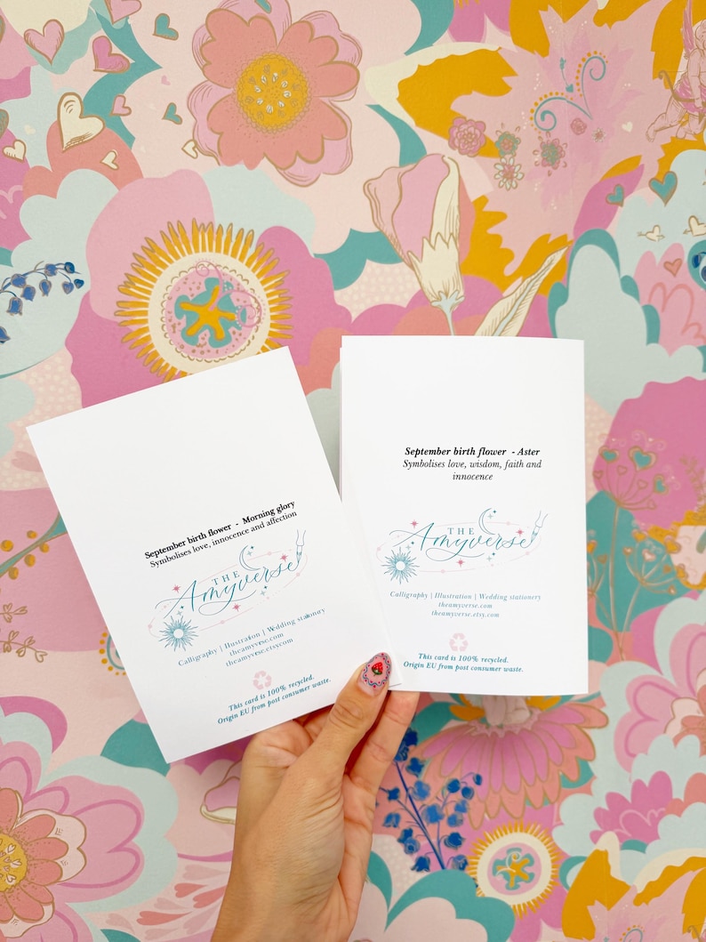 May include: Two white rectangular cards featuring black text and a blue logo. The left card reads September birth flower - Morning glory. The right card reads September birth flower - Aster. Both include The Amyverse branding and recycled paper info.