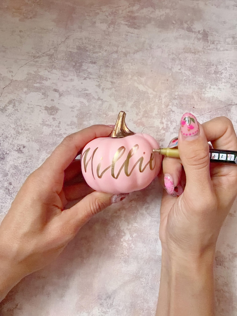 Pu&ograve; includere: Una piccola zucca decorativa rosa chiaro, liscia, con un picciolo color oro metallizzato. Il nome Millie &egrave; scritto in eleganti lettere dorate in corsivo sulla parte anteriore della zucca. Un pennarello a vernice dorata viene usato per scrivere.