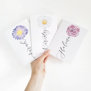 May include: Three rectangular white cards featuring watercolor flower illustrations and black cursive calligraphy. The first card shows a purple daisy with the name Sarah, the second a white daisy with the name Kirsty, and the third a pink rose with the name Helena.