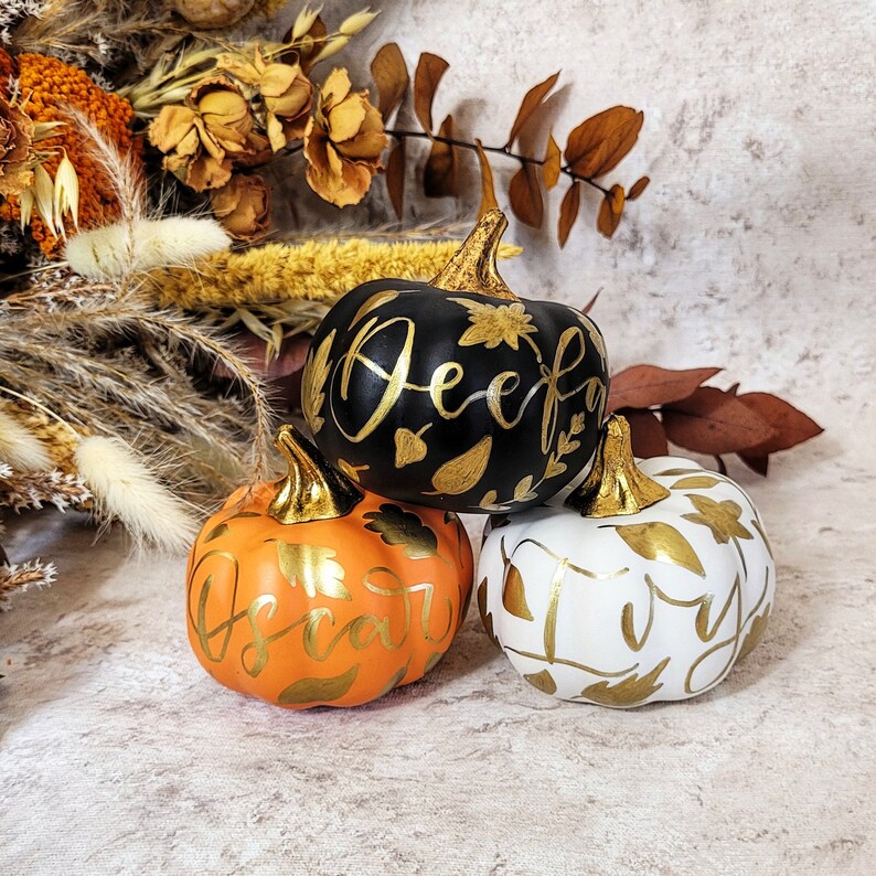 Pu&ograve; includere: Tre zucche decorative in nero, arancione e bianco, ognuna con un gambo dorato metallizzato e nomi scritti a mano in oro, tra cui Deefa, Oscar e Ivy. Le zucche sono decorate con motivi a foglia d'oro dipinti sulle loro superfici.