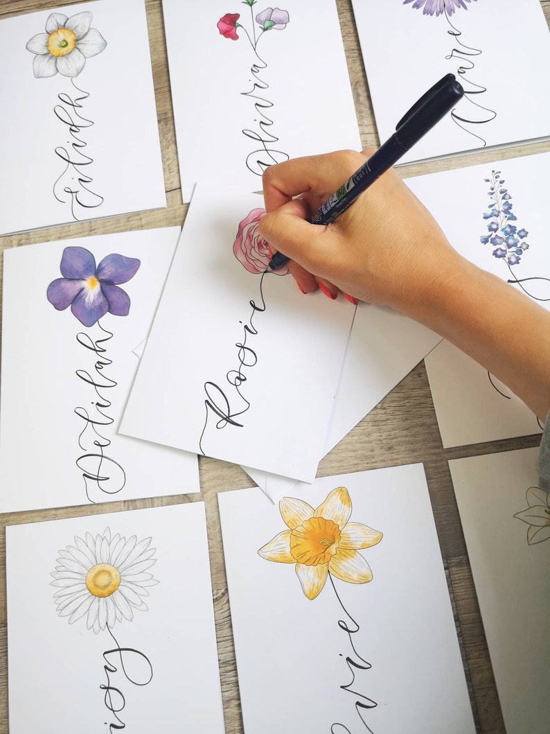 May include: A collection of white rectangular cards featuring hand-drawn watercolor flowers and elegant black cursive calligraphy names. Each card displays a unique floral illustration, including a daisy, daffodil, and rose, paired with a name.