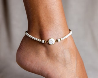May include: Ankle bracelet featuring small white beads and a central gold-toned charm with a white spiral design. The bracelet is worn on an ankle, showcasing a delicate and elegant accessory.