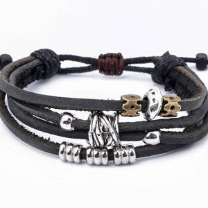 May include: A multi-strand black leather bracelet with silver and brass-toned metal accents. The bracelet features a central decorative element and adjustable black cord closure. The bracelet is a stylish accessory.