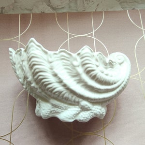 May include: A white ceramic seashell-shaped bowl with a textured surface. The bowl is on a pink and gold patterned background.