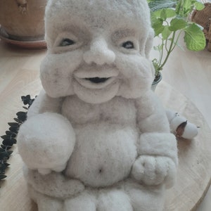 Peut inclure: Une sculpture en feutre blanc d'un bébé Bouddha souriant assis en position de méditation, les mains jointes devant la poitrine.