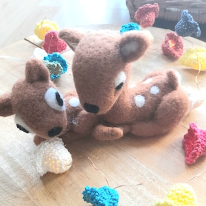 Peut inclure: Deux peluches de cerfs marrons et blancs tachetés se blottissent l'un contre l'autre. Les peluches sont en feutre et ont des yeux blancs. Les peluches sont entourées d'une guirlande de fleurs crochetées colorées.