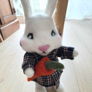Może przedstawiać: Biały królik z filcu w kratkowanej koszuli trzymający marchewkę. Królik ma różowe policzki i czarne oczy.