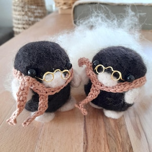 May include: Two handcrafted felted figures, each wearing a black cap, gold wire-rimmed spectacles, and a crocheted scarf. The figures have black bead eyes and are mainly black and white, with tan scarves.