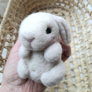 Peut inclure: Un petit lapin blanc en feutre aiguillé avec des yeux noirs et un nez rose. Le lapin est assis dans la main d'une personne.