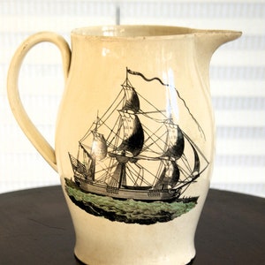 May include: A cream-coloured ceramic jug with a black and white illustration of a sailing ship. The ship has a large sail and is surrounded by waves.