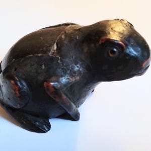 May include: A dark brown frog ornament with red detailing on the eyes and feet. The frog is depicted in a seated posture, with its legs folded beneath. The ornament has a textured finish and seems to be crafted from wood or a comparable material.