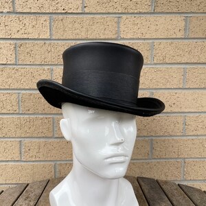 May include: A black top hat with a textured band and a slightly curved brim. The hat is displayed on a white mannequin head, set against a brick wall and a wooden table.