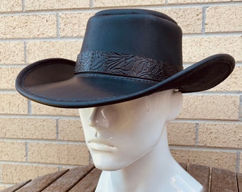 May include: A black leather cowboy hat with a decorative embossed band. The hat has a wide brim and a rounded crown. The hat is displayed on a white mannequin head.