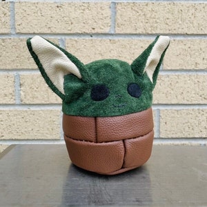 May include: A green and brown character doll with large ears and black eyes. The doll's head is green with tan ears, and the body is brown. The doll is sitting on a metal surface in front of a brick wall.