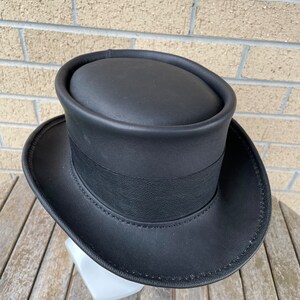 May include: A black top hat with a wide brim and a black band around the crown. The hat is made of a smooth, possibly leather-like material. The hat is sitting on a white stand.