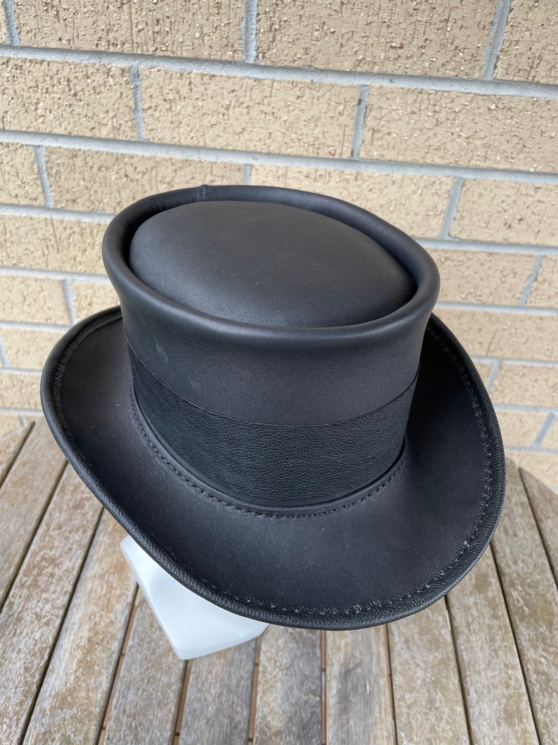 May include: A black top hat with a wide brim and a black band around the crown. The hat is made of a smooth, possibly leather-like material. The hat is sitting on a white stand.