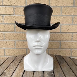 May include: A black top hat with a textured band and brim, displayed on a white mannequin head. The hat is set against a brick wall and a wooden table.