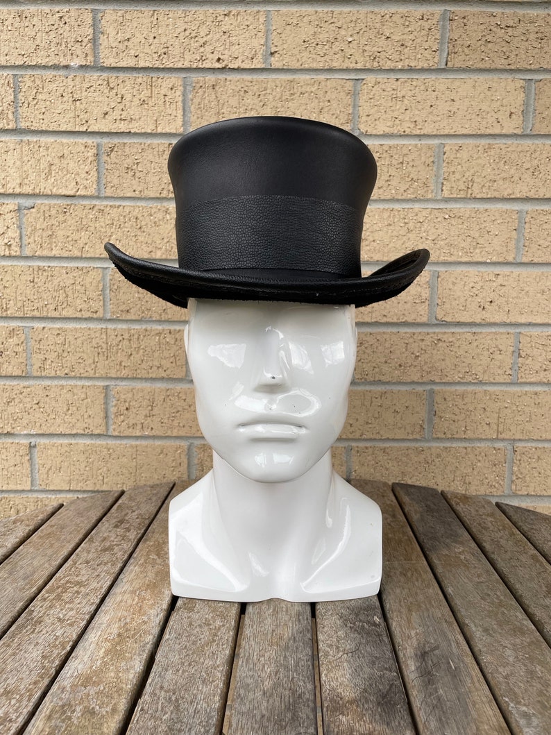 May include: A black top hat with a textured band and brim, displayed on a white mannequin head. The hat is set against a brick wall and a wooden table.