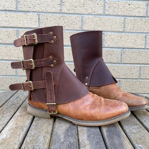 May include: A pair of brown leather boots with a distressed finish. The boots feature a mid-calf height with a wrap-around leather panel secured by three buckles. The boots are set on a wooden surface.