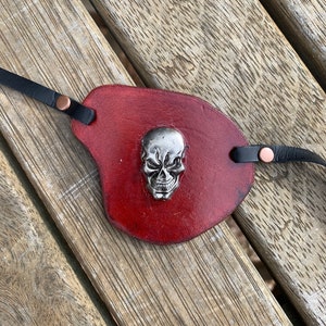 May include: A burgundy leather eye patch featuring a silver skull emblem. It has black straps with copper rivets. The eye patch is designed to cover one eye.