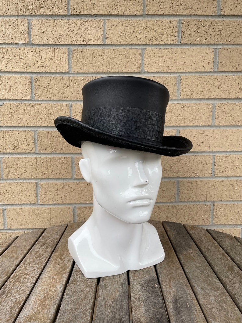 May include: A black top hat with a textured band and a slightly curved brim. The hat is displayed on a white mannequin head, set against a brick wall and a wooden table.