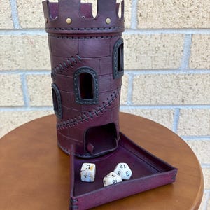 May include: A burgundy leather dice tower with a matching tray. The tower has a castle design with stitched details and small windows. Three dice are in the tray. The tower is on a wooden surface.