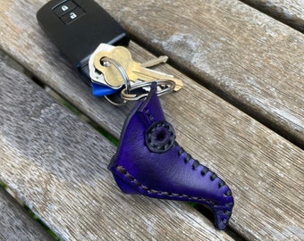 May include: A handmade, purple leather plague doctor mask keychain. The keychain is next to a car key fob and keys on a wooden surface. The mask features black stitching and a circular eye detail, showcasing a unique, gothic accessory.