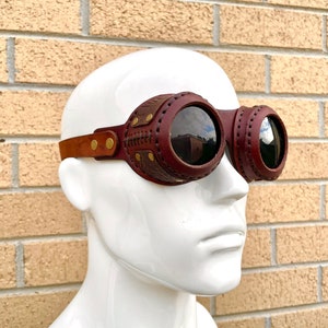 May include: Steampunk-style goggles with dark red leather frames and lenses. The goggles have a brown leather strap and are adorned with brass-colored rivets. The lenses are round and reflect the surroundings. The goggles are displayed on a white mannequin head.