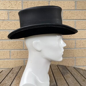 May include: A black top hat with a slightly curved brim and a textured band. The hat is displayed on a white mannequin head against a brick wall and wooden table.
