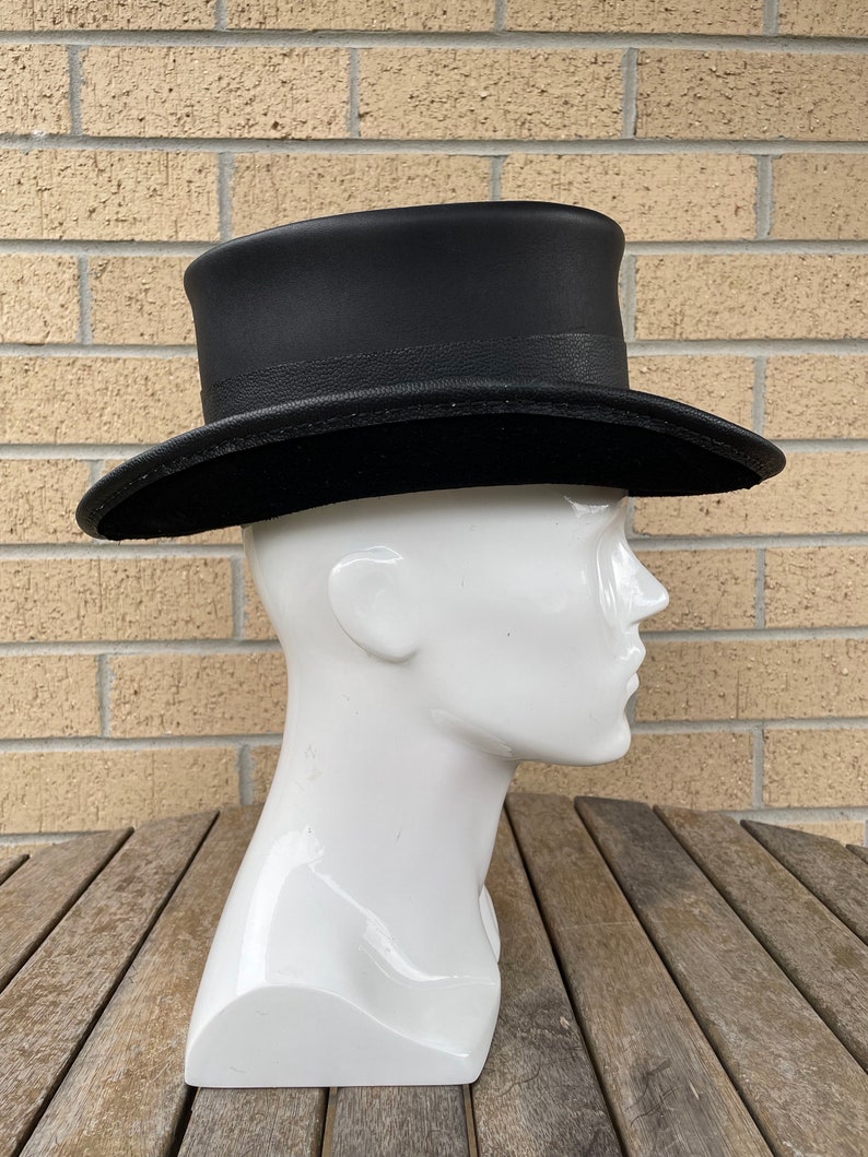 May include: A black top hat with a slightly curved brim and a textured band. The hat is displayed on a white mannequin head against a brick wall and wooden table.