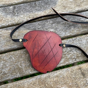 May include: A handmade, reddish-brown leather eye patch with a textured surface and a black strap. The eye patch is secured with small brass rivets and features a unique, carved design. The strap is designed to be worn around the head.