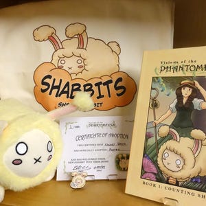 Puede incluir: Un juguete de peluche amarillo con forma de oveja con un certificado de adopción para un "Shabbit" de la serie de cómics "Visions of the Phantomfield". El certificado está numerado 1/1000 e indica que "Eurel Weich" ha adoptado oficialmente a "Pietro".