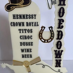 Op de afbeelding: Een crèmekleurig acrylbord in de vorm van een cowboylaars met de woorden "Hennessy", "Crown Royal", "Titos", "Ciroc", "Dusse", "Wine" en "Beer". Een hoefijzer en een bord met de tekst "The Hoedown" zijn ook inbegrepen.