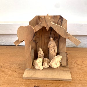 May include: A wooden nativity scene with a star on top, a manger with figures of Mary, Joseph, and the baby Jesus, and two sheep. The scene is made of olive wood and has a rustic, hand-crafted look.