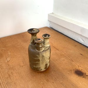 May include: A ceramic vessel featuring a rounded base and three distinct, narrow necks of varying heights. The piece is finished with a mottled glaze in shades of earthy brown and creamy beige, creating a textured appearance across the surface.