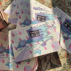 Könnte beinhalten: Eine Stapel von Kunstdrucken mit einem Retro-Computer-Design. Die Drucke zeigen den Text "GIVE UP?" und "YES" auf dem Bildschirm. Das Kunstwerk verwendet ein Pastellfarbschema mit rosa Tropfenformen und einem Gamecontroller.