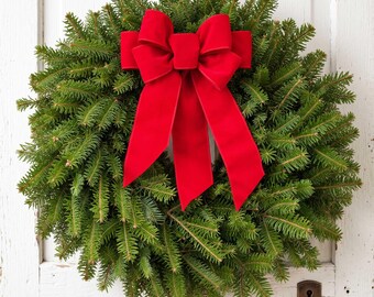 May include: A circular wreath made of dense green evergreen needles featuring a large, vibrant red velvet bow centered at the top. The bow has two prominent loops and two long, flowing tails that drape downward over the center of the foliage.