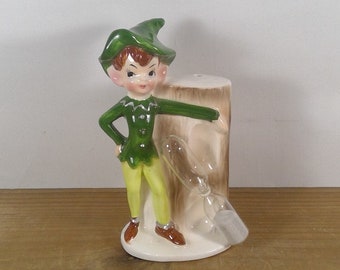 May include: A green and yellow ceramic figurine of an elf standing next to a tree stump with an hourglass inside. The elf is wearing a green hat and a green shirt with a white trim. The elf is holding onto the tree stump with one hand.