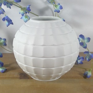 May include: White ceramic vase with a geometric pattern. The vase is round and has a wide opening. It is sitting on a wooden surface with blue flowers.