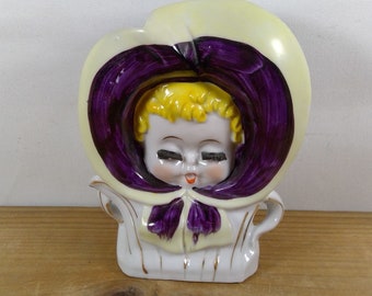 May include: A vintage ceramic figurine of a child's face framed by a large, light yellow bonnet with a deep purple interior. The child has yellow hair, closed eyes, and rosy cheeks. The white base is decorated with gold accents.