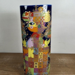 May include: A tall, cylindrical vase with a dark blue rim and a colorful, abstract design. The artwork features a stylized figure with geometric patterns in shades of yellow, orange, green, and purple, accented with gold details. The vase is likely made of ceramic.
