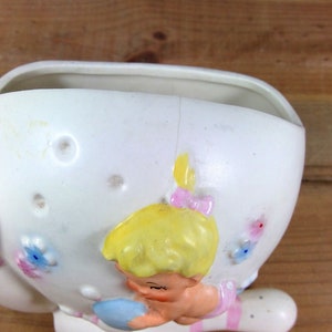 May include: White ceramic planter shaped like a girl holding a bowling pin. The girl has blonde hair, pink shoes, and a blue dress. The planter has pink and white stripes on the bowling pin and floral details.