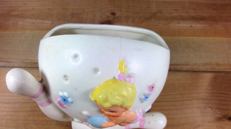 May include: White ceramic planter shaped like a girl holding a bowling pin. The girl has blonde hair, pink shoes, and a blue dress. The planter has pink and white stripes on the bowling pin and floral details.