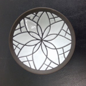May include: A small, round ceramic dish with a white glaze and a black geometric design. The design features a stylised flower with multiple petals.