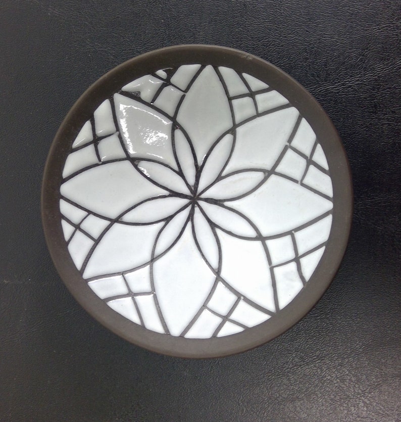 May include: A small, round ceramic dish with a white glaze and a black geometric design. The design features a stylised flower with multiple petals.