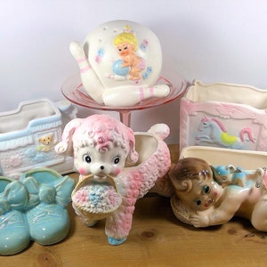 May include: A collection of vintage ceramic planters in pastel colors, featuring a baby in a crib, a lamb carrying a basket of flowers, a baby in a diaper, a bowling pin, and a pair of baby shoes.