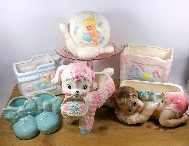 May include: A collection of vintage ceramic planters in pastel colors, featuring a baby in a crib, a lamb carrying a basket of flowers, a baby in a diaper, a bowling pin, and a pair of baby shoes.