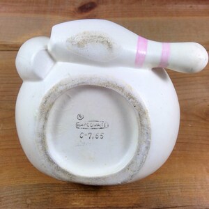 May include: White ceramic bowling pin shaped piggy bank with a pink stripe. The bottom of the bank has the text "(R) NAPCOWARE C-7765".